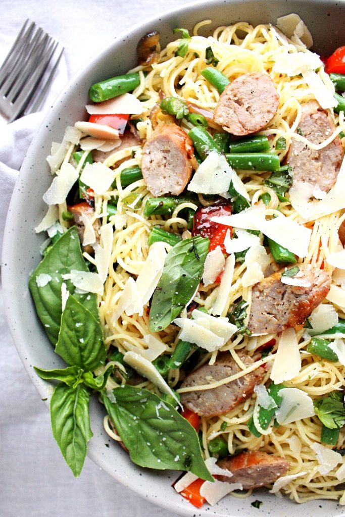 A bowl of grilled Italian sausage pasta with garden beans, fresh basil, red peppers, and a lemon dressing.
