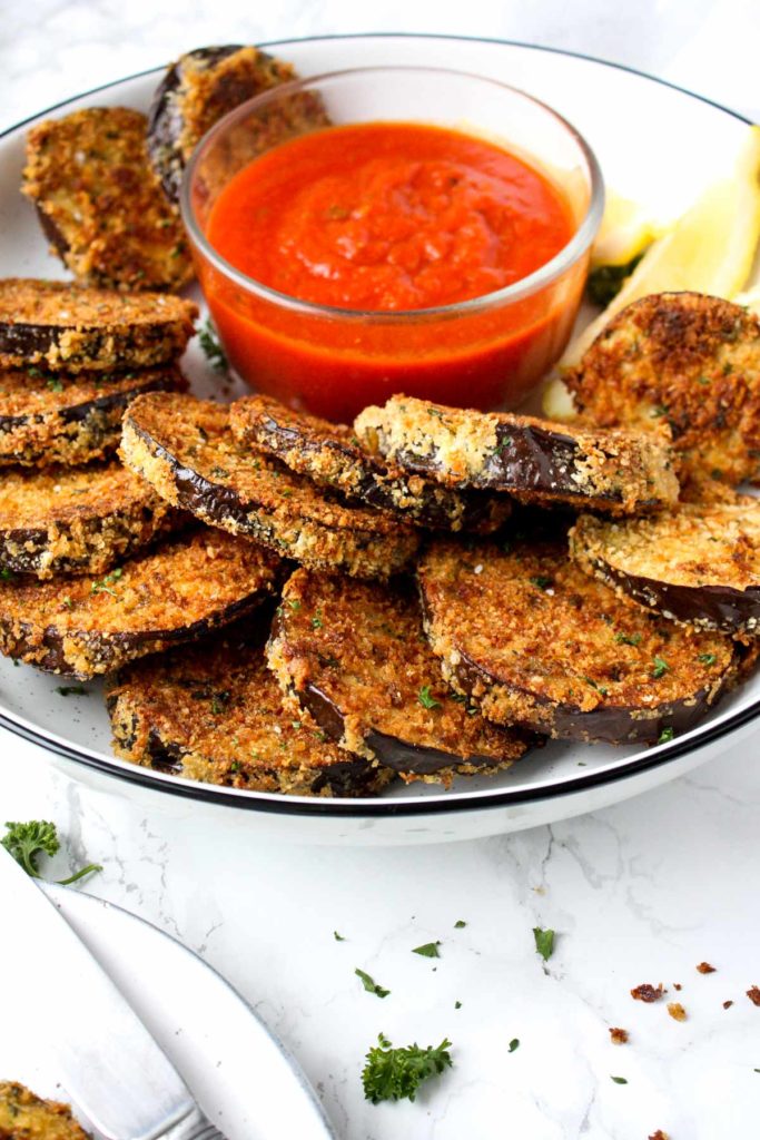 A platter of crispy baked eggplant rounds with a small bowl of marinara dipping sauce and lemon wedges.