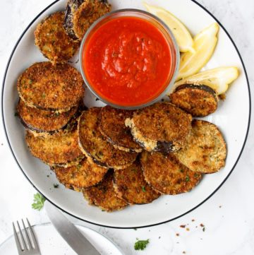 A platter of crispy baked eggplant rounds with a small bowl of marinara dipping sauce and lemon wedges.