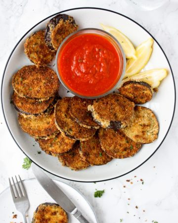 A platter of crispy baked eggplant rounds with a small bowl of marinara dipping sauce and lemon wedges.