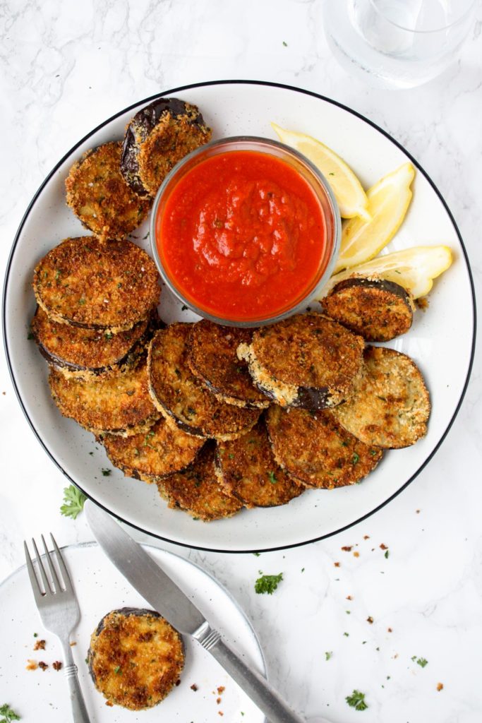 A platter of crispy baked eggplant rounds with a small bowl of marinara dipping sauce and lemon wedges.