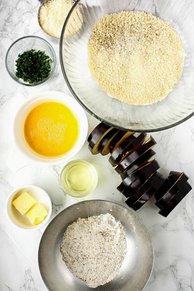 Ingredients ready for making baked eggplant: sliced eggplant, flour, panko, parmesan, parsley, eggs, butter, and oil.