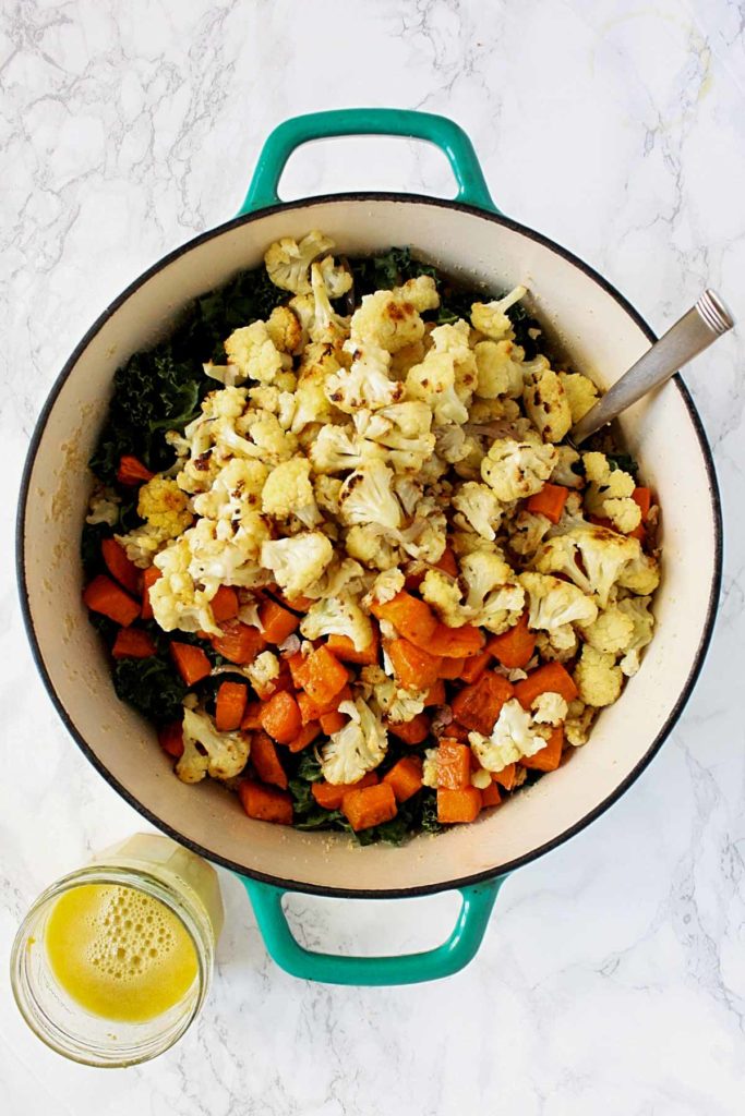 Pot of couscous topped with roasted sweet potatoes and cauliflower and a citrus dressing in a mason jar beside the pot.