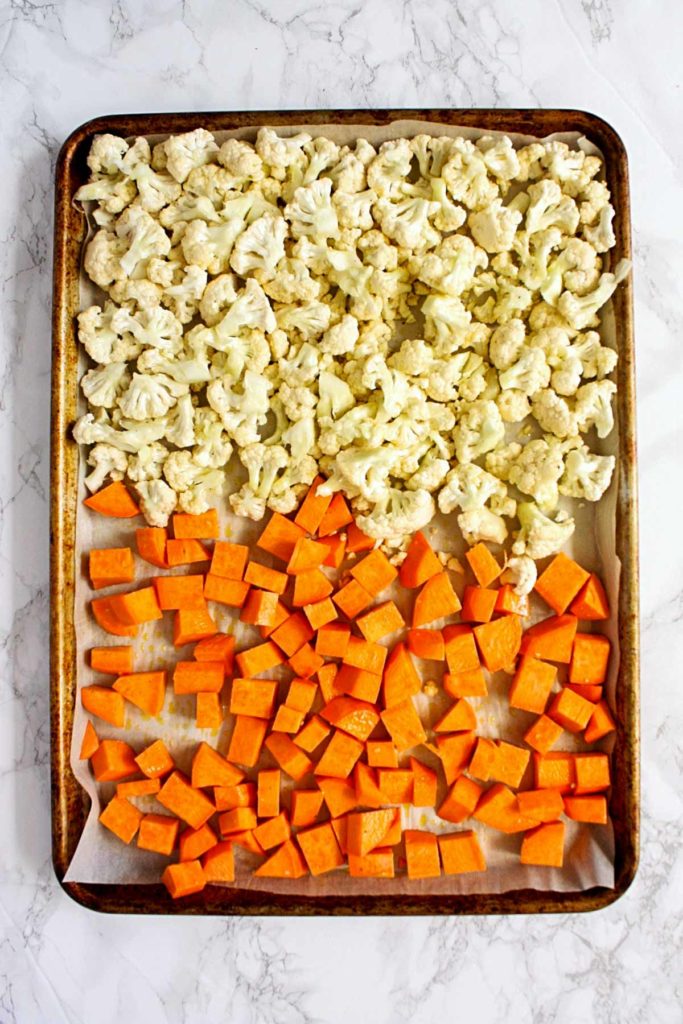 Chopped sweet potatoes and cauliflower florets on a parchment lined baking sheet, ready for roasting.