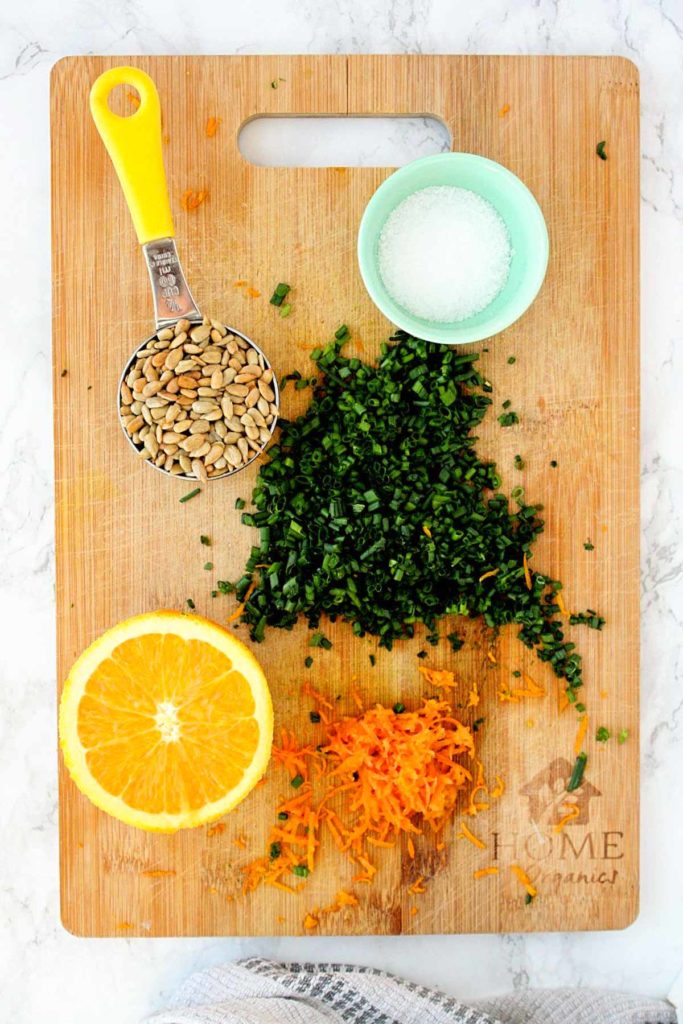 Couscous salad ingredients on a wooden cutting board: grated orange zest, sunflower seeds, chopped chives, and kosher salt.